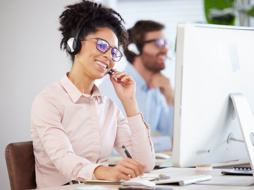 Smiling woman with headset and glasses at a desk on a call, male coworker in background, bright office setting.