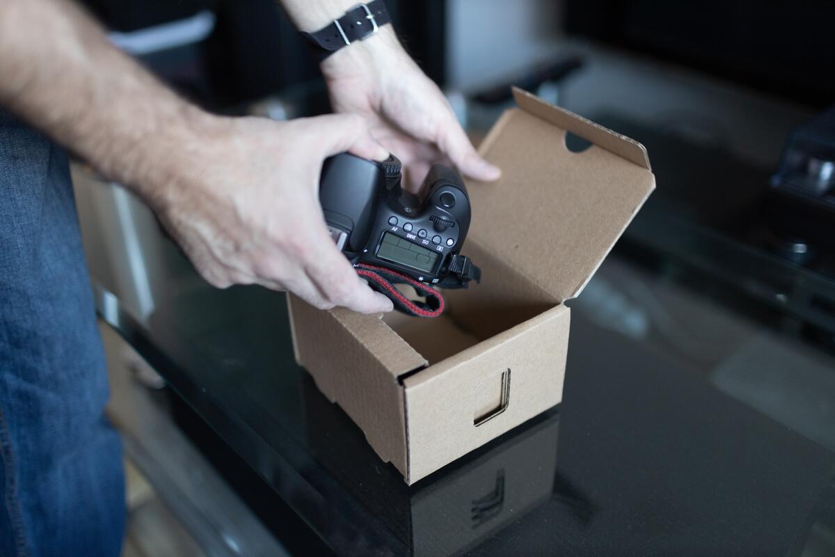 Hands carefully placing a camera into an open cardboard box on a glass table, conveying care and organization.